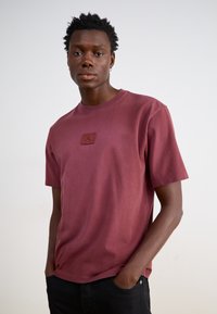 Mørkerød t-shirt med korte ærmer lavet af blødt stof, der har et lille struktureret logo patch på brystet. Casual pasform med rund halsudskæring.