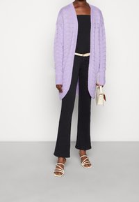 Lila gestrickte Strickjacke mit strukturiertem Muster, offener Vorderseite, langen Ärmeln, getragen über einem schwarzen Oberteil und dunklen Schlaghosen, kombiniert mit Riemchensandalen.