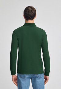 Grünes Langarm-Polohemd mit Kragen. Weiches Material, taillierte Passform und gerippte Bündchen. Kombiniert mit hellblauen Jeans.