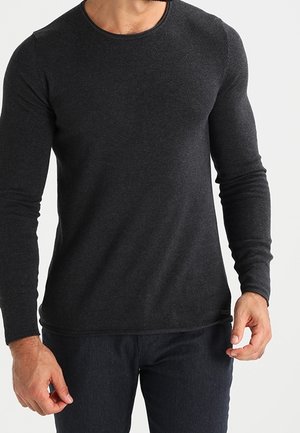 Man die een nauwsluitend donkergrijs shirt met lange mouwen en een donkere broek draagt, staand met de armen ontspannen langs zijn zij.