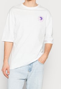 Camiseta de algodón blanca con mangas cortas, que presenta un gráfico de yin-yang morado rodeado de rayos en el lado izquierdo del pecho. Diseño de cuello redondo estándar.