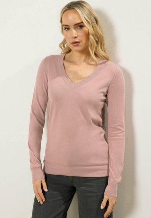 Blondynka w jasnoróżowym swetrze z dekoltem w szpic i ciemnych dżinsach, stojąca na tle białej ściany, patrząca przed siebie z neutralnym wyrazem twarzy.