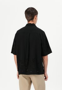 Μαύρο κοντομάνικο πουκάμισο με κομμένα λουλουδάτα σχέδια στο κάτω άκρο. Το πουκάμισο έχει γιακά και άνετη εφαρμογή.