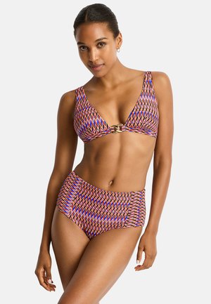 Mujer con bikini de talle alto con patrón geométrico colorido y cierre metálico en la parte superior, posando frente a un fondo liso.