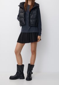 Schwarze gepolsterte Weste mit Kapuze, graues Langarmshirt, schwarzer plissierter Rock und schwarze Leder-Ankle Boots mit Schnallen-Detail.