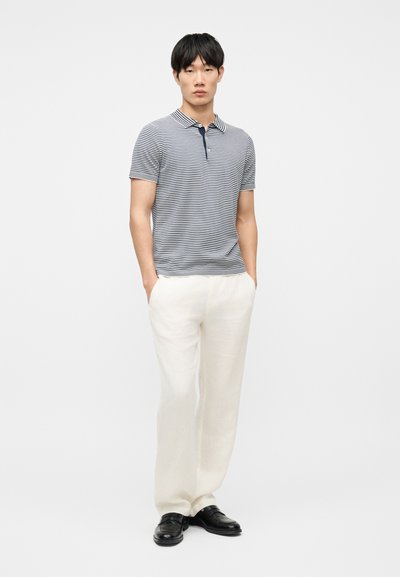 Homme debout, les mains dans les poches, portant un polo rayé bleu marine et blanc, un pantalon blanc et des chaussures noires, sur un fond blanc.