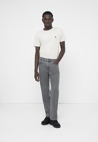Jeans en denim gris avec une coupe droite, associés à un T-shirt beige clair arborant un petit logo coloré. Des chaussures noires complètent le look.