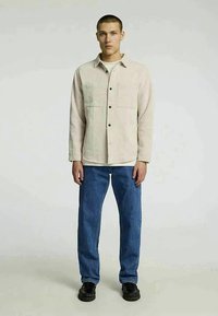 Gaiši bēša pogu krekls ar divām priekšējām kabatām, apvienots ar zilu džinsu biksēm un melniem apaviem. Gluda tekstūra, brīvs piegriezums un minimāls dizains.