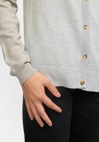 Svetlo szedý cardigan vyrobený z mäkkej pleteniny s zlatými gombíkmi a ribovanými manžetami. Nosený cez tmavú látku, predvádza priliehavý strih.