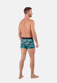 Men's swim trunks featuring a floral pattern in turquoise, with a black waistband, fitted design, and mid-thigh length. Textured fabric.