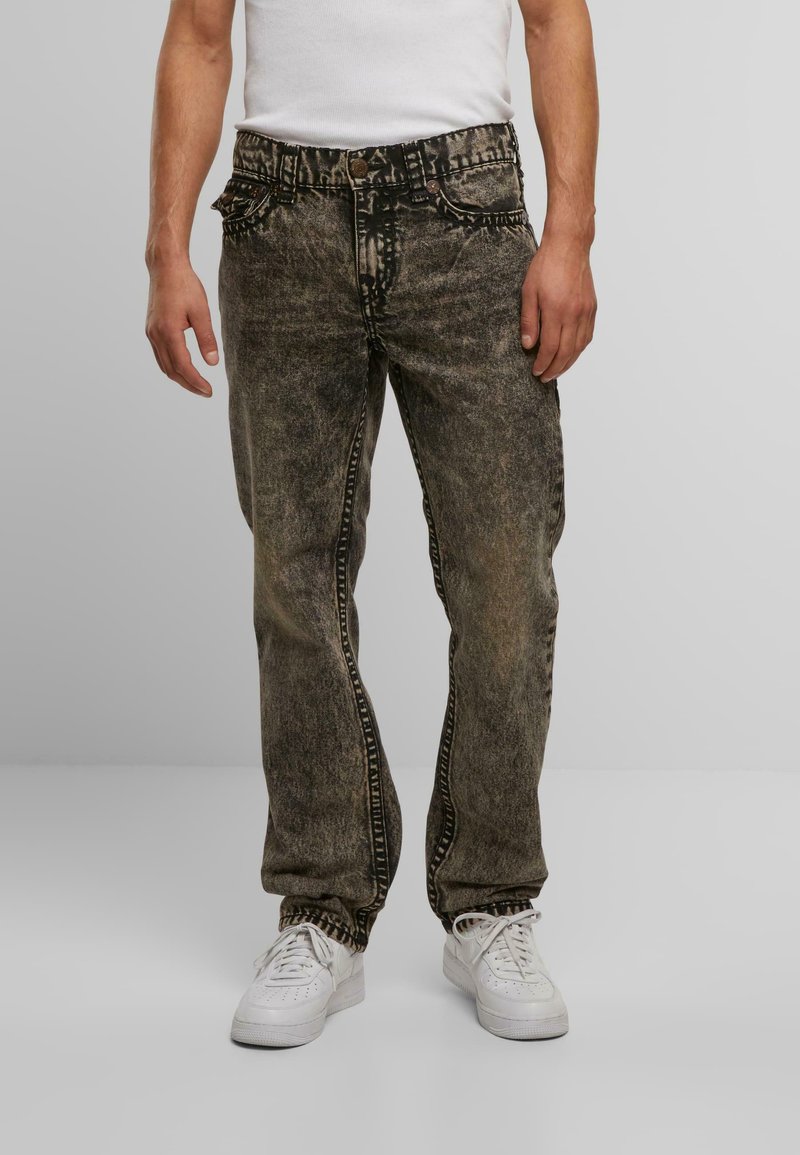 Jeans de mezclilla negra desgastada con un corte relajado, que presentan detalles de costura distintos y una textura sutilmente desgastada. Combinados con zapatillas blancas.