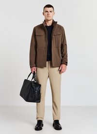 Veste marron avec deux poches poitrine, haut bleu marine en dessous, pantalon beige et sac en cuir noir. Des chaussures noires complètent la tenue.