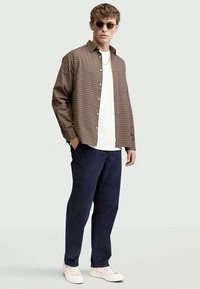 Καφέ καρό πουκάμισο με κουμπιά, λευκό μπλουζάκι, ναυτική παντελόνι και λευκά αθλητικά παπούτσια. Μοντέλο φορά στρογγυλά γυαλιά ηλίου και λουρίδα.