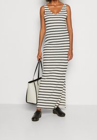 Robe maxi tricotée rayée en noir et blanc avec un décolleté rond. Le modèle tient un sac fourre-tout noir et blanc et porte des sabots noirs avec des clous.