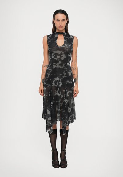 Femme tatouée portant une robe sans manches en camouflage noir et gris avec un décolleté à trou, une jupe mi-longue transparente, des chaussettes noires jusqu'aux genoux et des talons noirs.