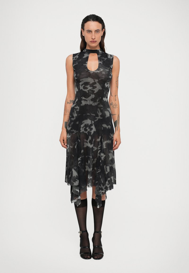 Vrouw met tatoeages draagt een mouwloze zwart-grijze camouflage jurk met een keyhole halslijn, een doorschijnende jurk tot halverwege de knie, zwarte kniekousen en zwarte hakken.