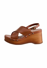 Sandaletta con zeppa in pelle marrone, con cinture incrociate, fibbia regolabile alla caviglia, suola effetto legno e borchie decorative in metallo.