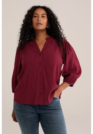 Kvinde med langt krøllet hår iført øreringe i ringform, en løs mørkebrun bluse med pufærmer og blå jeans, stående med den ene hånd i lommen.