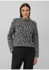 Vrouw in een grijs en zwart luipaardprint trui en zwarte broek, staand met één hand in de zak tegen een effen achtergrond.