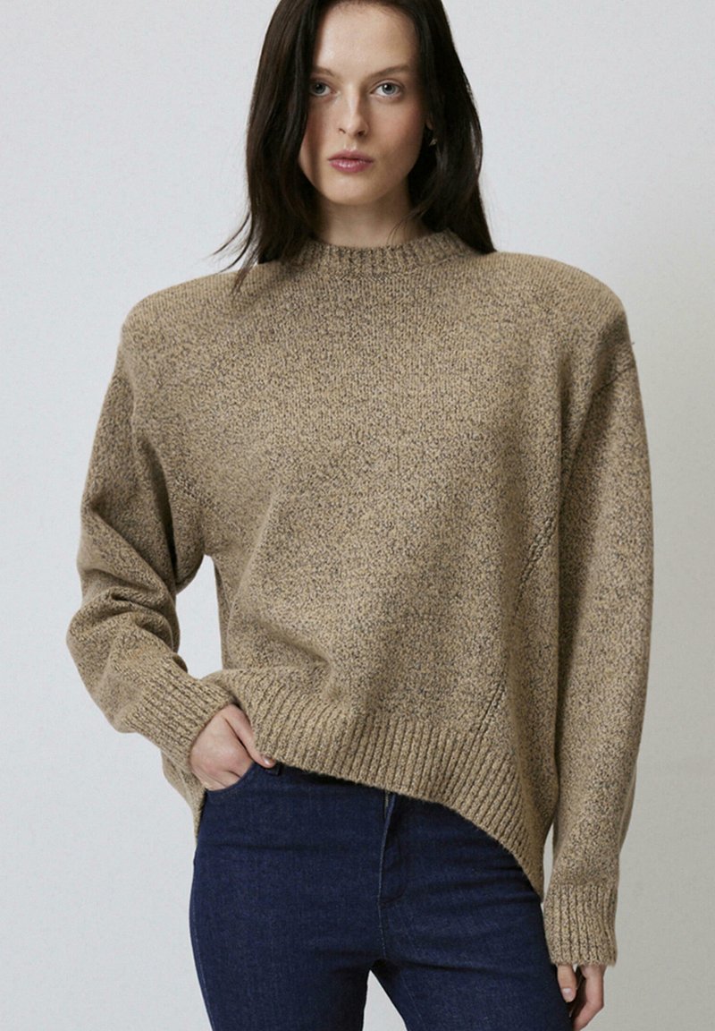 Maglione beige a trama con collo alto, polsini e orlo a coste, e un taglio leggermente asimmetrico. Abbinato a jeans blu scuro.