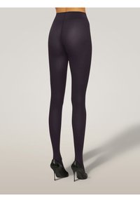 Persona che indossa collant opachi di un viola scuro e scarpe col tacco nere, vista da dietro su uno sfondo neutro.