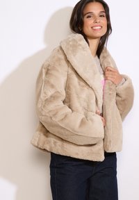 Beige faux fur jacket met een brede kraag, cropped ontwerp en zachte textuur. Gekleed over een lichte trui en gecombineerd met donkere jeans.