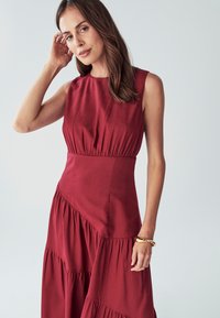 Rdeča obleka brez rokavov s tesno prilegajočim zgornjim delom in plastmi volančkov na robu. Narejena iz mehkega, teksturiranega materiala. Preprost, eleganten dizajn.