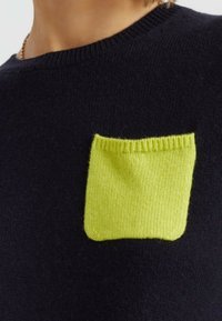 Navy gebreide trui met een contrasterende felgroene vierkante zak met ribbelrand, die textuur en een schoon ontwerp toont.