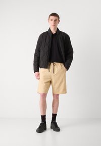 Black quilted bomber jacket, black polo shirt, beige textured shorts, black sneakers, black socks with red logo. Simple casual outfit.