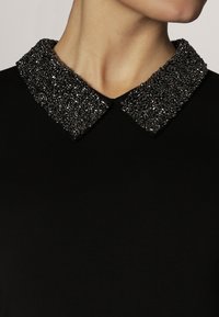 Kvinna som bär en svart topp med en bred, detaljerad Peter Pan-krage dekorerad med glänsande svarta pärlor.