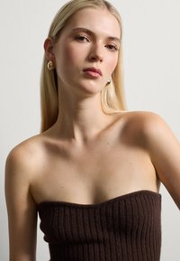 Top senza spalline a coste marrone con un design aderente. Pelle liscia di tonalità naturale, capelli biondi lunghi e dritti, e orecchini metallici rotondi.