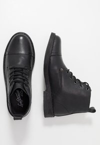 Botas de cuero negro hasta el tobillo con punta redondeada, diseño de cordones, suela de goma plana y ligeros detalles de costura en los costados.
