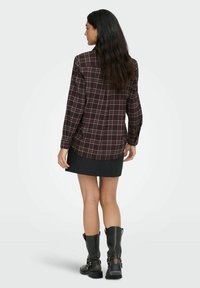 Kvinde med langt, mørkt hår iført en brunternet skjorte, mørk miniskirt og sorte støvler til midt på læggen, stående med ryggen til mod en hvid baggrund.