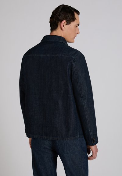 Mężczyzna odwrócony plecami, ubrany w ciemnoniebieską kurtkę dżinsową i pasujące dżinsy na jasnym, jednolitym tle.