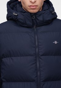 Homme portant une doudoune bleu marine avec un col montant zippé et une capuche réglable, affichant le logo GANT sur la poitrine.