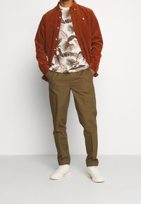 Eine rostfarbene Corduroy-Hemdjacke über einem weißen T-Shirt mit Palmblattdruck, kombiniert mit olivgrünen Hosen und weißen Turnschuhen.