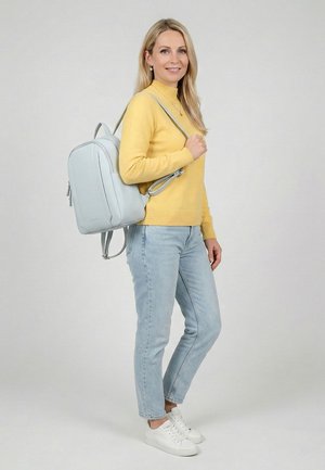 Žena s dlouhými blond vlasy, oblečená ve žlutém svetru, světle modrých džínách, bílých teniskách a nesoucí na rameni světle šedý batoh.