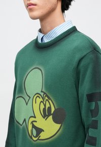 Zöld pulóver nagy sárga Mickey egér grafikával elöl. Texturált anyagból készült, bordázott nyakkivágással. Fekete felirat a ujján.