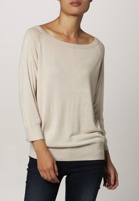 Beige pletený svetr s širokým výstřihem, tříčtvrtečními rukávy a žebrovanými manžetami. Měkká textura s uvolněným střihem, nošený přes tmavé džíny.