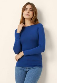 Bluză albastră, cu mâneci lungi, ajustată, dintr-un material moale și elastic, cu decolteu rotund și textură netedă, asortată cu blugi albaștri deschiși.