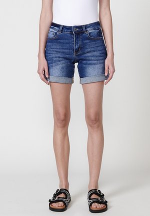 Denim-Shorts in einem mittleren Blauton, mit umgeschlagenen Säumen, fünf Taschen und einem Knopfverschluss-Design. Kombiniert mit schwarz-weißen Sandalen.