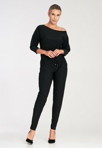 Czarny kombinezon z dekoltem odsłaniającym ramiona, trzyczwartowymi rękawami, podkreśloną talią i zwężanymi nogawkami. Wykonany z gładkiego, elastycznego materiału.