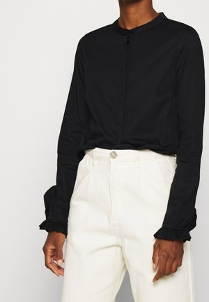 Person trägt eine langärmelige schwarze Bluse mit Rüschen an den Ärmeln und eine weiß Hochbundhose, steht vor einem einfarbigen Hintergrund.