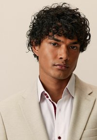 Jachetă de costume bej cu model herringbone, cu rever ascuțit, purtată peste o cămașă albă cu butoane și accente roșii închise. Păr creț de culoare închisă.