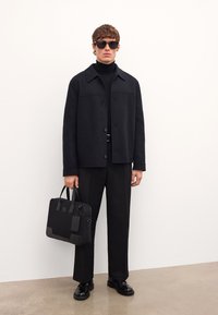 Musta villatakki, poolopaita ja housut, joissa on aurinkolasit. Pitelee mustaa rakennerikasta nahkalaukkua, jossa on logo ja tekstuuriset yksityiskohdat.