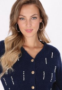 Cárdigan de punto azul marino con cuello en V, embellecido con acentos parecidos a perlas y botones dorados a lo largo del frente. Textura suave, diseño ajustado.