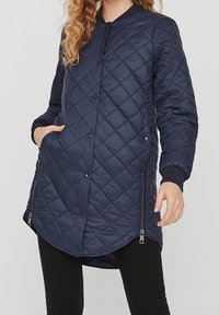 Marinblå quiltad lång jacka med tryckknappar, dragkedjefickor, ribbade muddar och rundad fåll. Tillverkad av lätt, isolerat material.