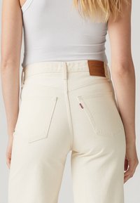 Mujer con jeans Levi’s de talle alto color blanco hueso y una camiseta sin mangas blanca de canalé, vista desde atrás, desde la cintura hasta la parte superior de los muslos.