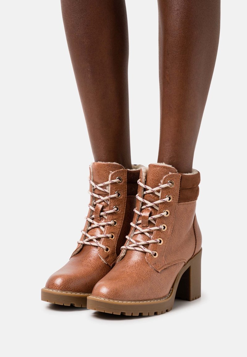 Bottines en cuir marron à lacets, talons épais et col rembourré portées sur des jambes à la peau foncée, sur un fond uni.