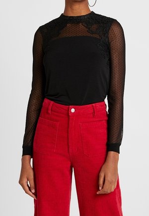 Black long-sleeve sheer lace top with polka dot pattern paired with high-waisted bright red corduroy pants with front pockets.